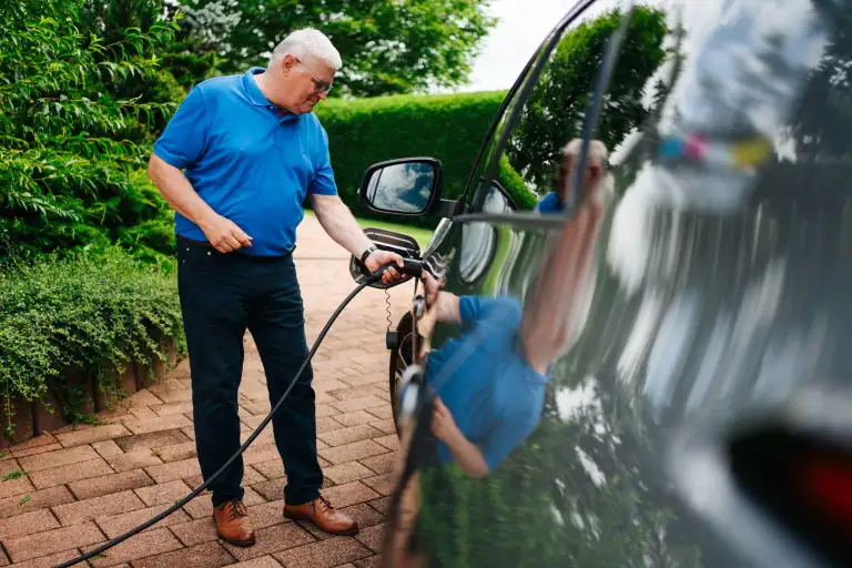 Mann lädt Elektroauto auf Einfahrt vor grüner Hecke auf.